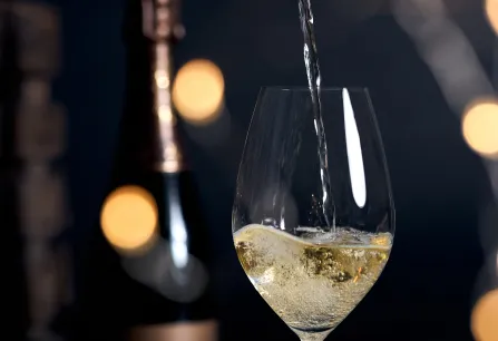 Weißwein wird in elegantes Glas gegossen, im Hintergrund unscharfe Lichter und Flasche.