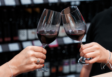 Zwei Personen stoßen mit Rotweingläsern an, Weinregale im Hintergrund.