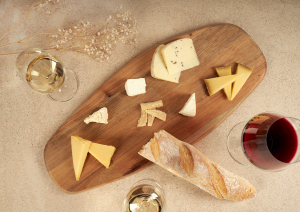 Holzbrett mit verschiedenen Käsesorten, Baguette und Weingläsern auf heller Oberfläche.