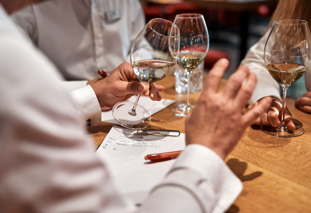 Personen in weißen Hemden diskutieren bei einem Glas Weißwein an einem Holztisch.