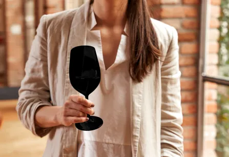 Frau in beigem Blazer hält schwarzes Weinglas vor Ziegelwand.