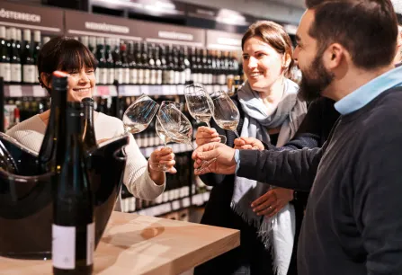 Gruppe hebt Weingläser, lächelnd in Weinladen, Flaschen im Vordergrund.