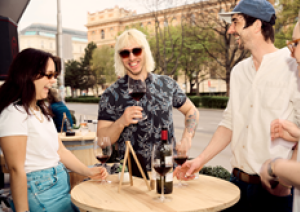 Gruppe von Freunden lacht und trinkt Wein an einem Holzstehtisch im Freien.