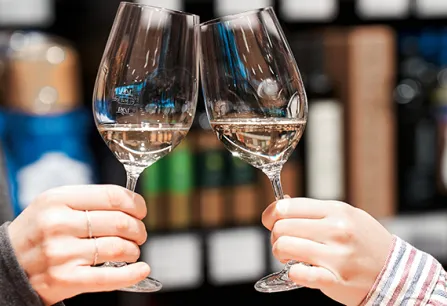 Zwei Personen stoßen mit gefüllten Weißweingläsern in einem Geschäft an.