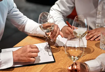 Personen in weißen Hemden halten Weingläser und schreiben Notizen auf eine Holzoberfläche.