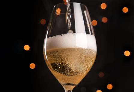 Glas Sekt wird eingeschenkt, schäumend, vor unscharfem, stimmungsvollem Hintergrund mit Lichtern.