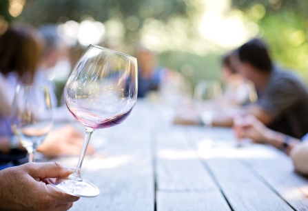 Weinglas auf Holztisch, Menschen im Hintergrund bei einem Freilufttreffen.