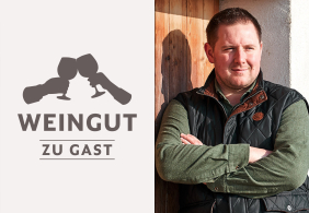Mann in grüner Hemdjacke, schwarz gestepptem Gilet, lehnt an Holzwand.
