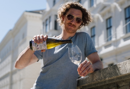 Mann mit Sonnenbrille schenkt Weißwein in Glas, städtische Kulisse im Hintergrund.