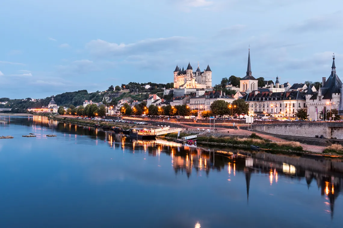 Historische Stadtansicht mit Fluss, beleuchteten Gebäuden und malerischem Schloss im Hintergrund.