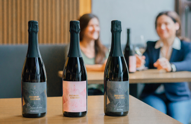 Drei Sektflaschen auf einem Tisch, Personen im Hintergrund beim Weingenuss in einem Café.