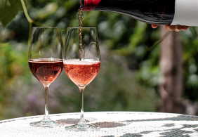 Zwei gefüllte Weingläser auf Tisch im Garten, Flasche im Hintergrund.