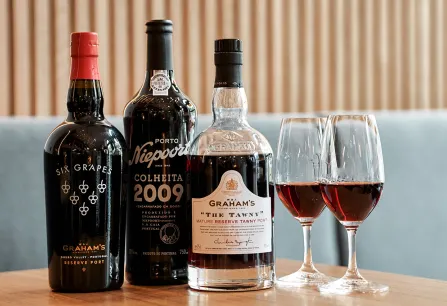 Drei Portweinflaschen und zwei Gläser auf Holztisch vor blauer Polsterbank.