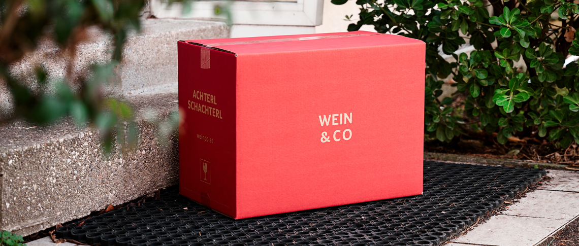Rote Versandbox auf schwarzer Fußmatte neben Treppenstufen und Sträuchern.