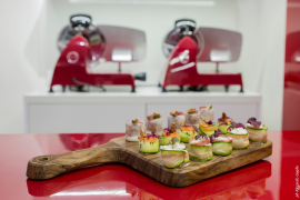 Holzbrett mit bunten, dekorativen Häppchen vor roten Aufschnittmaschinen auf roter Oberfläche.