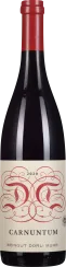 Die Weinflasche zeigt ein elegantes, geschwungenes Etikett mit roten Verzierungen und schwarzer Schrift. Der Flaschenkorken ist ebenfalls rot, passend zu den Etikett-Details. Die Flasche hat eine klassische Form mit dunkel getöntem Glas.