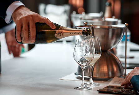 Eine Hand schenkt Sekt in elegante Gläser auf einer weißen Tischplatte ein.