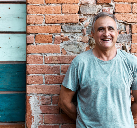 Mann in grauem T-Shirt lehnt an gemauerter Backsteinwand neben blauer Holztür.