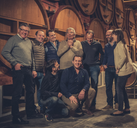 Gruppe von acht Menschen in einer Weinkellerei vor großen Holzfässern.