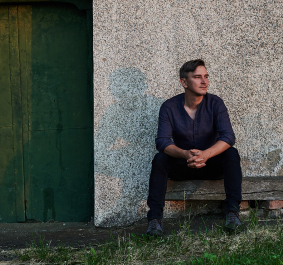 Mann sitzt auf Holzbank vor grüner Tür und rustikaler Hauswand.