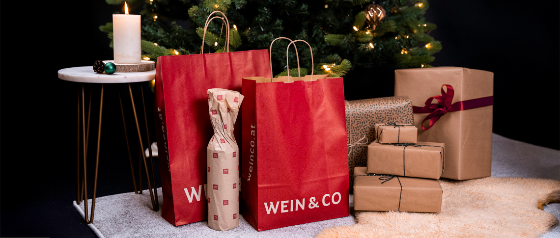 Rote Geschenktüten und verpackte Geschenke vor beleuchtetem Weihnachtsbaum auf Teppich.