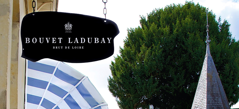 Schwarzes Schild mit Schriftzug, im Hintergrund ein Baum und ein Turm.