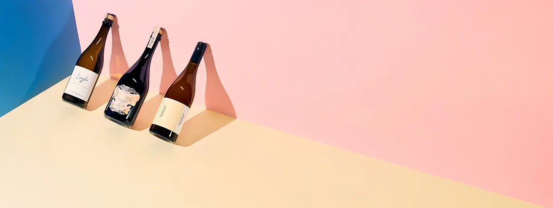 Drei Weinflaschen auf pastellfarbenem Hintergrund: Blau, Rosa und Beige.