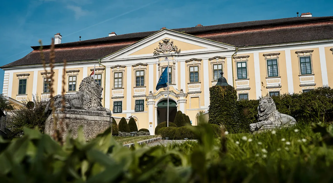 Gelbes Barockgebäude mit skulpturalen Löwen im Vordergrund und blauem Himmel.